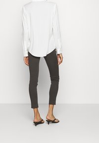 Camisa blanca de manga larga con dobladillo redondeado, combinada con pantalones gris ajustados y tacones abiertos negros, vista desde la espalda.