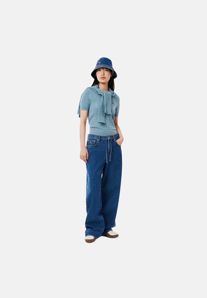 Top de punto azul claro con mangas cortas, acompañado de jeans de mezclilla azul de pierna ancha. Llevado con un sombrero de cubo azul y zapatos casuales.
