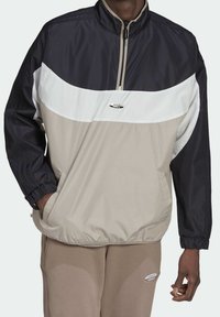 Person som bär en svart, vit och beige Adidas vindjacka med dragkedja, samt beige Adidas byxor med logotyper på båda plaggen.