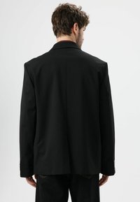 Zwarte op maat gemaakte blazer met gestructureerde schouders, enkele achterventilatie en twee knopen. Materiaal lijkt glad met minimale textuur.