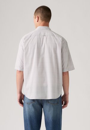 Mann trägt ein hellgraues kariertes Kurzarmhemd und blaue Jeans, von hinten vor einem einfarbigen Hintergrund.
