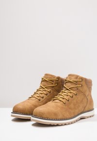 Botas de ante marrón hasta el tobillo con cordones amarillos, cuello acolchado y suela de goma blanca. Presentan costuras en contraste y un acabado texturizado.