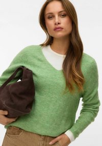 Femme aux longs cheveux bruns portant un pull vert clair à col en V et un pantalon beige, tenant un sac à main en suède marron foncé sur son côté gauche.
