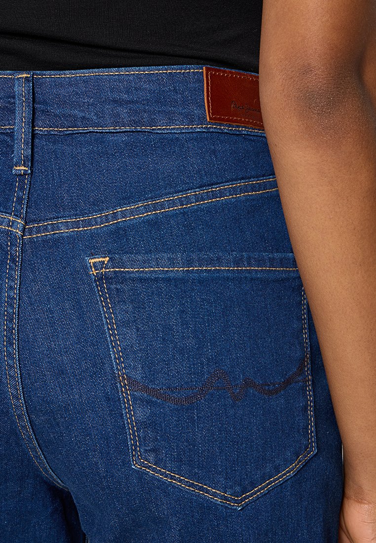 Jean en denim bleu foncé avec une étiquette de marque en cuir marron, des coutures visibles et un design unique sur la poche arrière.