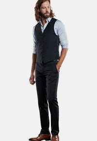 Dunkle Weste über einem hellblauen Hemd, kombiniert mit dunkler Hose und braunen Schuhen. Der Stoff wirkt glatt und maßgeschneidert.