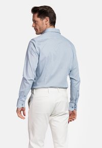 Chemise à manches longues bleu clair avec un petit motif géométrique, poignets boutonnés et col évasé, portée avec un pantalon blanc.