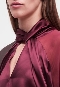 Γυναίκα που φοράει πουκάμισο από σατέν σε βαθύ μωβ χρώμα με ψηλό γιακά δεμένο σε κόμπο και λαιμόκοψη V, που αποκαλύπτει μέρος του λαιμού και των χειλιών της.