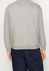 Persona con un maglione grigio chiaro a maniche lunghe e jeans blu scuro, vista da dietro con le mani rilassate lungo i fianchi.