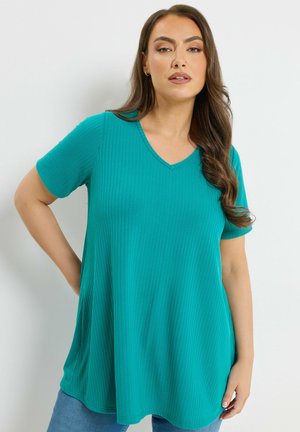 Frau mit langen braunen Haaren, die ein türkisfarbenes, geripptes Kurzarm-V-Ausschnitt-Oberteil und blaue Jeans trägt, steht vor einem weißen Hintergrund.