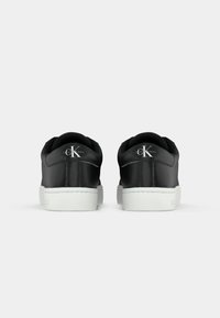 Pár čiernych tenisiek Calvin Klein s bielymi podrážkami, zobrazených zozadu, pričom je vidieť logo "cK Calvin Klein Jeans" na päte.