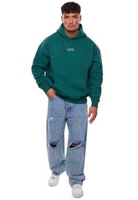 Sweatshirt à capuche teal avec une poche avant et un patch logo, associé à un jean bleu clair déchiré et des baskets blanches.