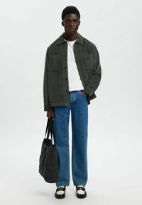 Giovane uomo che indossa una giacca bottonata verde scuro, una t-shirt bianca, jeans blu e scarpe bianco e nere, mentre tiene una borsa a tracolla grigia scura.