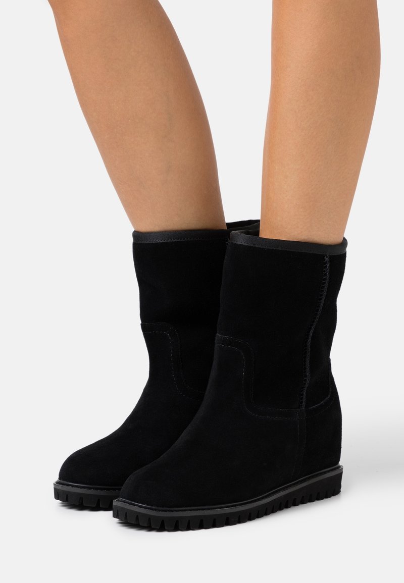 Bottes compensées mi-mollet en daim noir avec semelles en caoutchouc rainurées portées sur jambes nues sur fond clair uni.