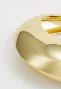 Anillo de metal dorado con una superficie lisa y reflectante, que presenta un diseño curvado y una sección abierta, destacando su textura pulida.