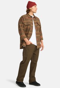 Koszula w kratę w ziemistych odcieniach, noszona na białym T-shircie, w zestawieniu z brązowymi spodniami i czarnymi trampkami. Czerwona czapka beanie na głowie.
