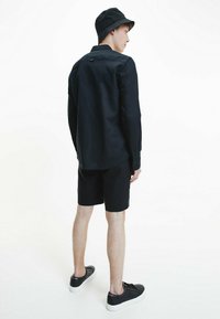 Camicia nera a maniche lunghe, pantaloni corti abbinati e un cappello a secchiello nero. Tessuto morbido, vestibilità comoda e sneakers nere con suola bianca. Design minimalista.