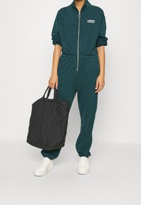 Dunkelgrüner Reißverschluss-Overall mit langen Ärmeln und elastischem Bund, kombiniert mit einer gesteppten schwarzen Tragetasche und weißen Plateauschuhen.