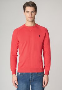 Maglione rosso lavorato a maglia con maniche lunghe, polsini e orlo a coste, caratterizzato da un piccolo logo scuro sul petto. Abbinato a jeans in denim blu.