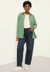 Green plaid flannel shirt over a beige top, paired with dark jeans and white sneakers. Features a relaxed fit and button details.