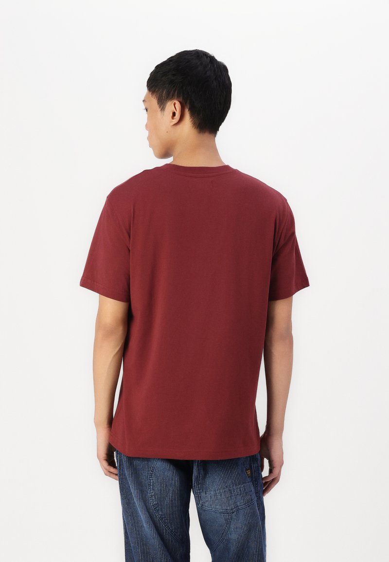 Bordeauxrotes Baumwoll-T-Shirt mit Rundhalsausschnitt und kurzen Ärmeln, das einen lässigen Schnitt und eine glatte Textur aufweist. Getragen mit blauen Streifenhosen.