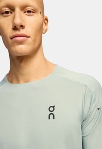 Jasnoniebieska sportowa koszulka z długim rękawem, okrągłym dekoltem i czarnym logo po lewej stronie na klatce piersiowej. Gładka faktura; minimalistyczny design.