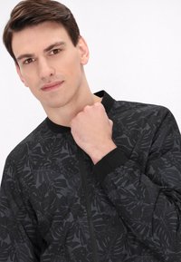 Czarna kurtka bomberka z teksturowanym wzorem liści, z prążkowanym kołnierzem i mankietami, zamkiem błyskawicznym z przodu oraz gładkim, lekkim materiałem.