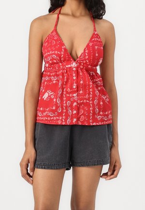 Žena v červeném halter topu s vzorem a předními knoflíky, v černých denimových šortkách, stojící před bílým pozadím.