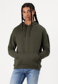 Olive green hooded sweatshirt with a kangaroo pocket, drawstring, and ribbed cuffs. Soft fabric and relaxed fit, paired with light blue jeans.