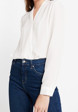Femme portant une blouse blanche à manches longues rentrée dans un jean bleu foncé taille haute, debout devant un fond blanc uni.