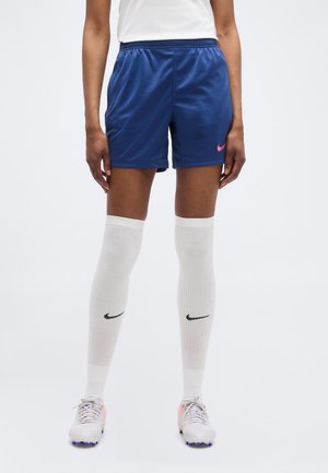 Άτομο που φορά μπλε αθλητικά σορτς, λευκές κάλτσες μέχρι το γόνατο με μαύρα λογότυπα, και λευκά παπούτσια ποδοσφαίρου με μπλε καρφιά, στέκεται μπροστά σε απλό φόντο.