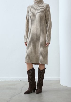 Vestito beige a coste in maglia con collo alto e maniche lunghe, abbinato a stivali da cowboy in suede marrone scuro con dettagli ricamati.