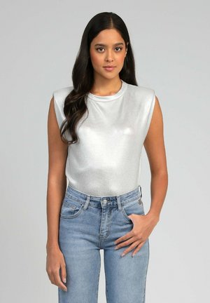 Femme aux longs cheveux foncés portant un haut argenté sans manches avec des épaulettes et un jean taille haute bleu clair, debout devant un fond uni.