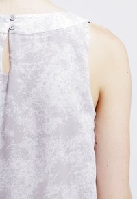 Blusa senza maniche grigia con un motivo testurizzato, chiusura a bottone sul retro e dettaglio a occhio di chiave sul collo.