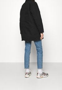 Sort frakke med lige snit, kombineret med lyseblå jeans og sølvfarvede sportssko. Synligt logo og snørebånd på skoene.