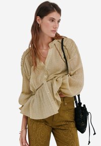 Blusa gialla chiara con maniche gathered e dettagli in pizzo, caratterizzata da una silhouette ampia. Abbinata a pantaloni verde oliva con ricami floreali.