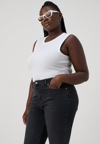 Hvit ribbet tanktop sammen med mørkegrå skinny jeans. Modellen har på seg hvite, vinklete solbriller og har fletter, med hendene i lommene.