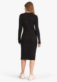 Robe noire à manches longues côtelées avec une silhouette ajustée, longueur genoux, et motif texturé, portée avec des baskets claires.