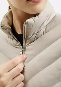 Jasno beżowa pikowana kurtka z zapięciem na zamek, z wysokim kołnierzem i ukośnymi wzorami przeszycia. Gładka faktura, zamek błyskawiczny do regulacji ręcznej.