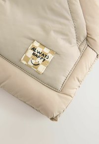Beżowa kurtka puchowa w rogu z teksturowanej tkaniny, pikowanym wzorem i kwadratową naszywką z napisem "ZAWSZE SZCZĘŚLIWY" oraz uśmiechniętą buźką.