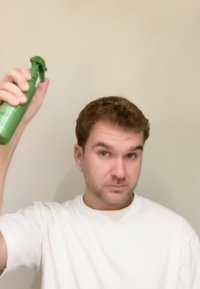 Man with short brown hair wearing white shirt sprays green bottle near his head against plain beige wall.