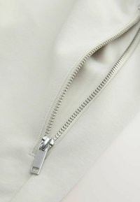 Gros plan d'une fermeture éclair en métal argenté partiellement ouverte sur un tissu beige clair, montrant les détails des dents et du curseur.