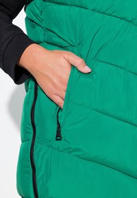 Gilet puffa verde con design trapuntato, dotato di una tasca con zip sul lato e una texture liscia. Accento nero all'ascella.