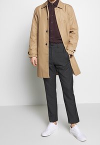 Trench beige sopra una camicia scozzese color bordeaux, abbinata a pantaloni grigi su misura e sneaker bianche. Tessuto liscio, taglio classico.
