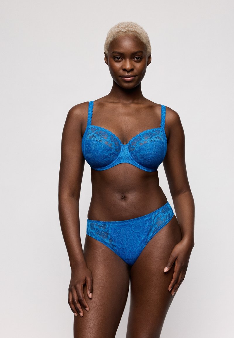 Femme aux cheveux blonds courts portant un soutien-gorge en dentelle bleue et une lingerie assortie, debout devant un fond clair uni.