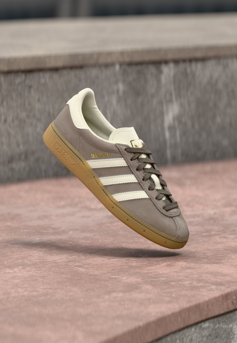 Ανοιχτό καφέ σουέντ αθλητικό παπούτσι Adidas με λευκές ρίγες και σόλα από καουτσούκ, αναρτημένο πάνω από ροζ πέτρινα σκαλοπάτια σε εξωτερικό χώρο.