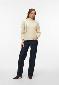 Krēmkrāsas adīts džemperis ar augstu apkakli un pelēkām detaļām, kombinēts ar tumšām taisna griezuma džinsa biksēm un brūnām, ar smailiem purngaliem kurpēm.