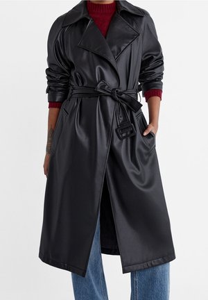 Personne portant un long trench-coat en cuir noir avec une ceinture nouée sur un pull rouge et un jean bleu, les mains dans les poches du manteau.