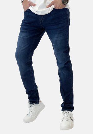 Jeans slim-fit blu scuro realizzati in denim, con una texture liscia e una leggera scoloritura, abbinati a sneakers bianche.