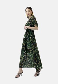 Durchsichtiges schwarzes Maxikleid mit grünem und gelbem Blumenmuster, tailliertem Bund und kurzen Ärmeln, kombiniert mit schwarzen Riemchenschuhen.