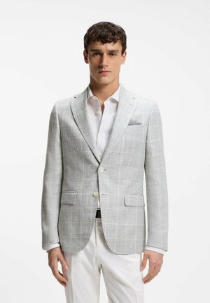 Uomo che indossa blazer a quadri grigio chiaro sopra camicia bianca e pantaloni bianchi, in piedi davanti a uno sfondo bianco uniforme.